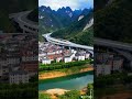 一条高速公路蜿蜒穿梭于巍峨的崇山峻岭之间，宛如银色巨龙盘旋于青翠山峦之上。高速公路下方，一座宁静的小县城依山傍水而建，红瓦白墙的房屋错落有致，炊烟袅袅升起，展现出一派祥和的生活气息。