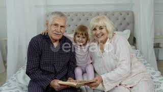 Cute Girl with Senior Retired Grandmother and Grandfather Sitting on Bed Reading Book in Bedroom...