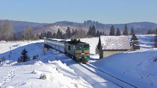 A passenger train descends into a sunny valley overlooked by snow-capped mountain peaks